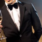 A man's upper body wearing a black tuxedo. He is holding a glass of champagne.