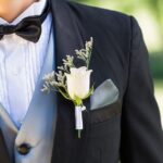 A man in a black tuxedo and gray waistcoat stands in a lush green garden, wearing a white rose boutonniere.
