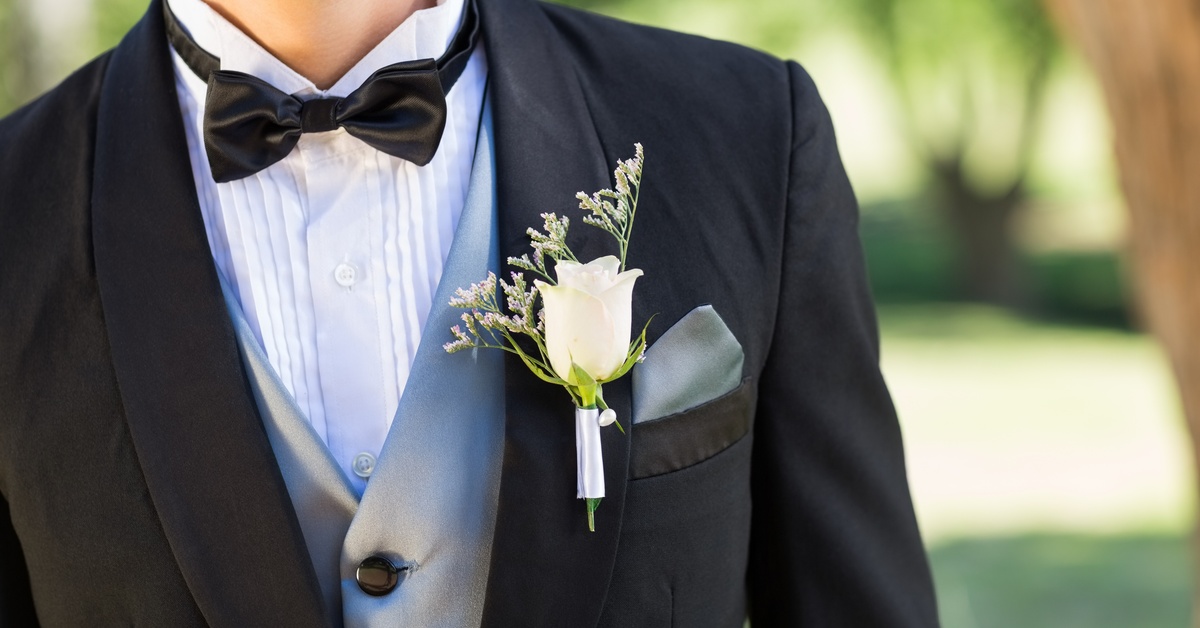 A man in a black tuxedo and gray waistcoat stands in a lush green garden, wearing a white rose boutonniere.