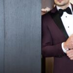 A man in a dark burgundy, custom-tailored tuxedo standing in a doorway against a dark-colored wooden accent wall.