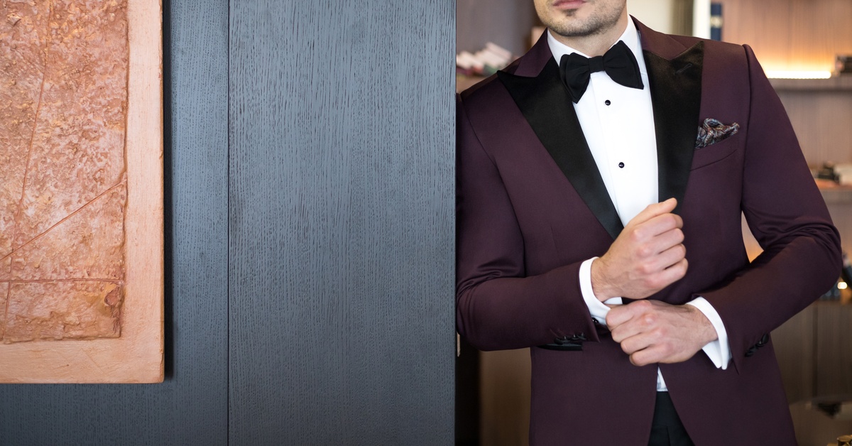 A man in a dark burgundy, custom-tailored tuxedo standing in a doorway against a dark-colored wooden accent wall.