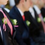 A row of sitting groomsmen wearing black tuxedos with pink ties, and a pinned white rose boutonniere.