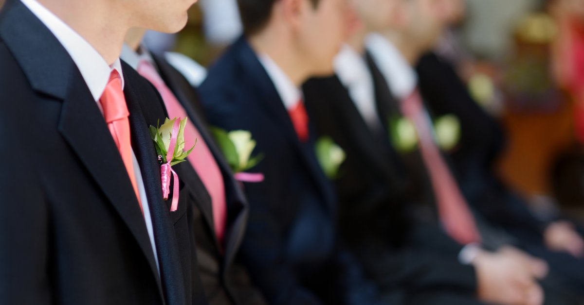 A row of sitting groomsmen wearing black tuxedos with pink ties, and a pinned white rose boutonniere.